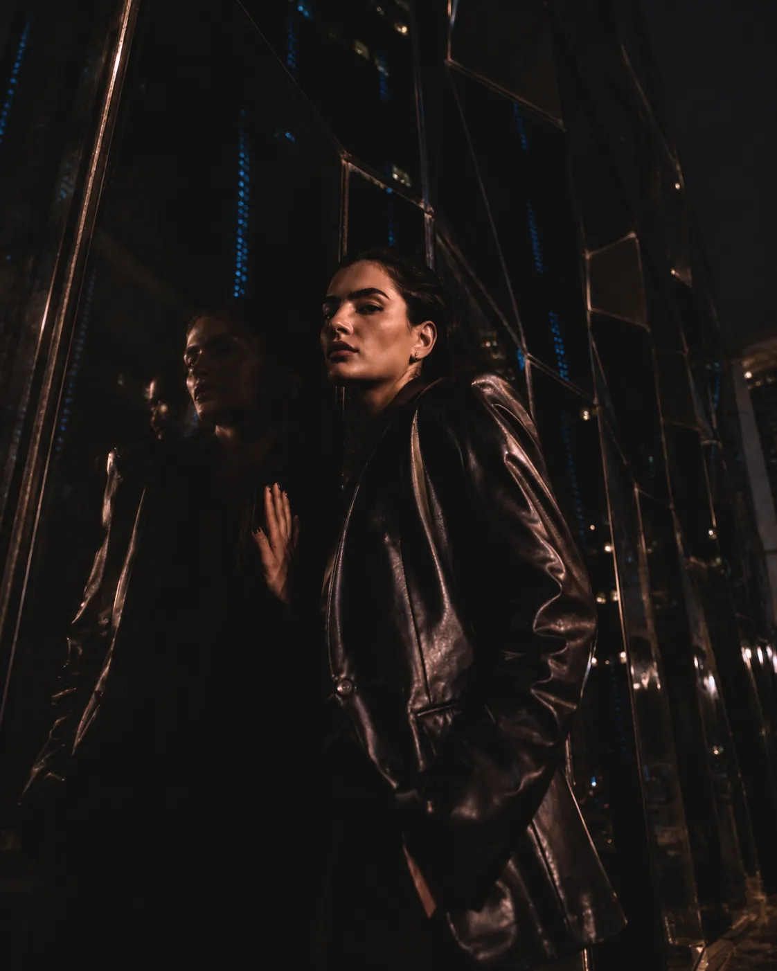 Fashion model in a dark leather jacket posing beside reflective glass at the base of the Jordan Gate Towers in Amman at night.
