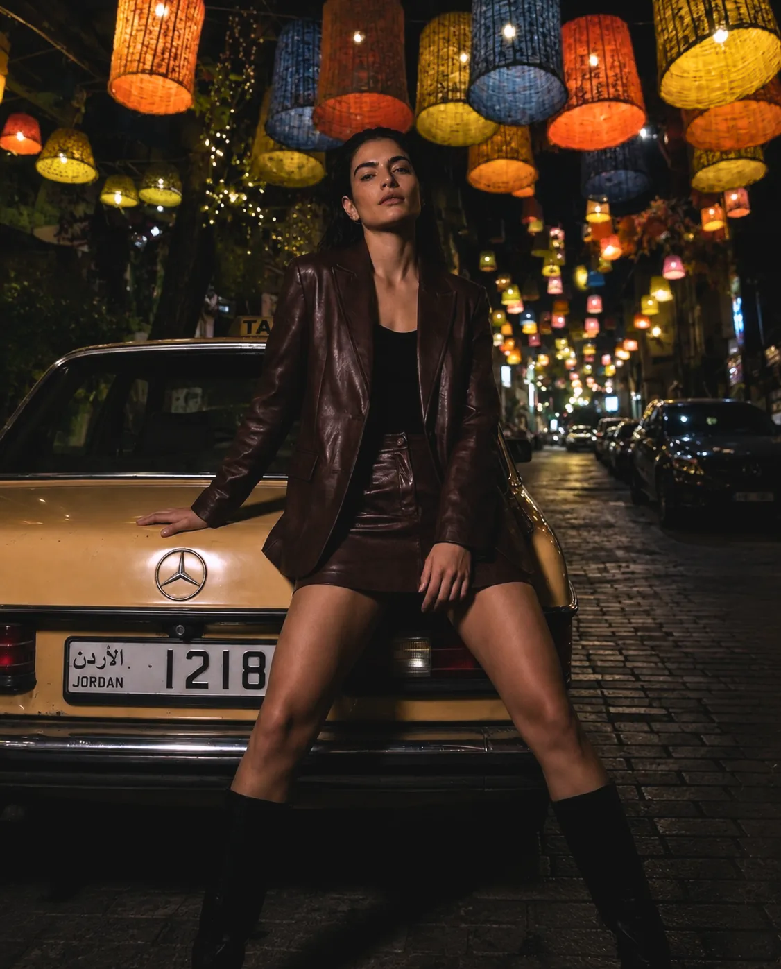 Fashion model in a dark red dress sitting at night on Rainbow Street in Amman with illuminated buildings and street lights behind her.