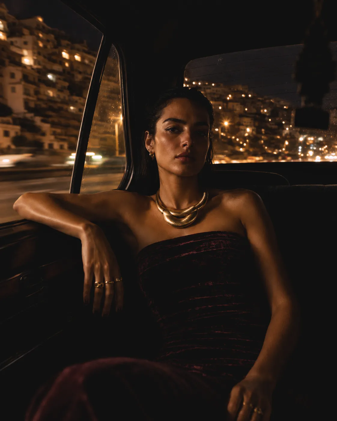 Fashion model in a burgundy leather outfit leaning against a cream Jordanian taxi on a busy Amman street beneath colourful hanging lights at night.