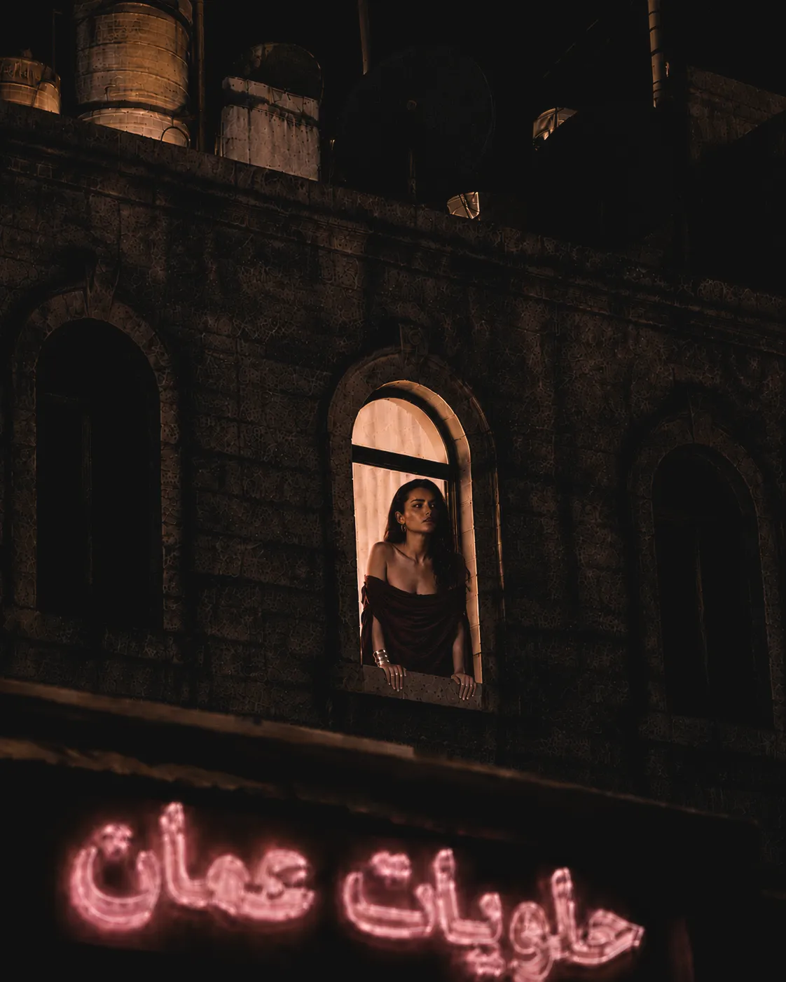 Fashion model framed in a stone window above a glowing Arabic neon sign on an Amman street at night.