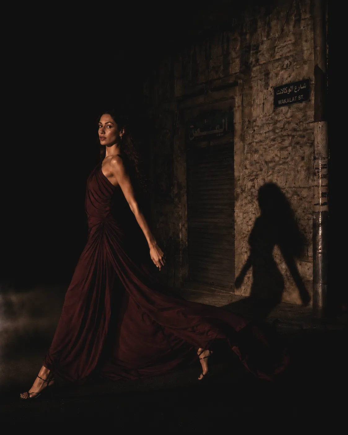 Fashion model in a flowing dark red gown walking along Wakalat Street in Amman at night beside stone buildings and shopfronts.