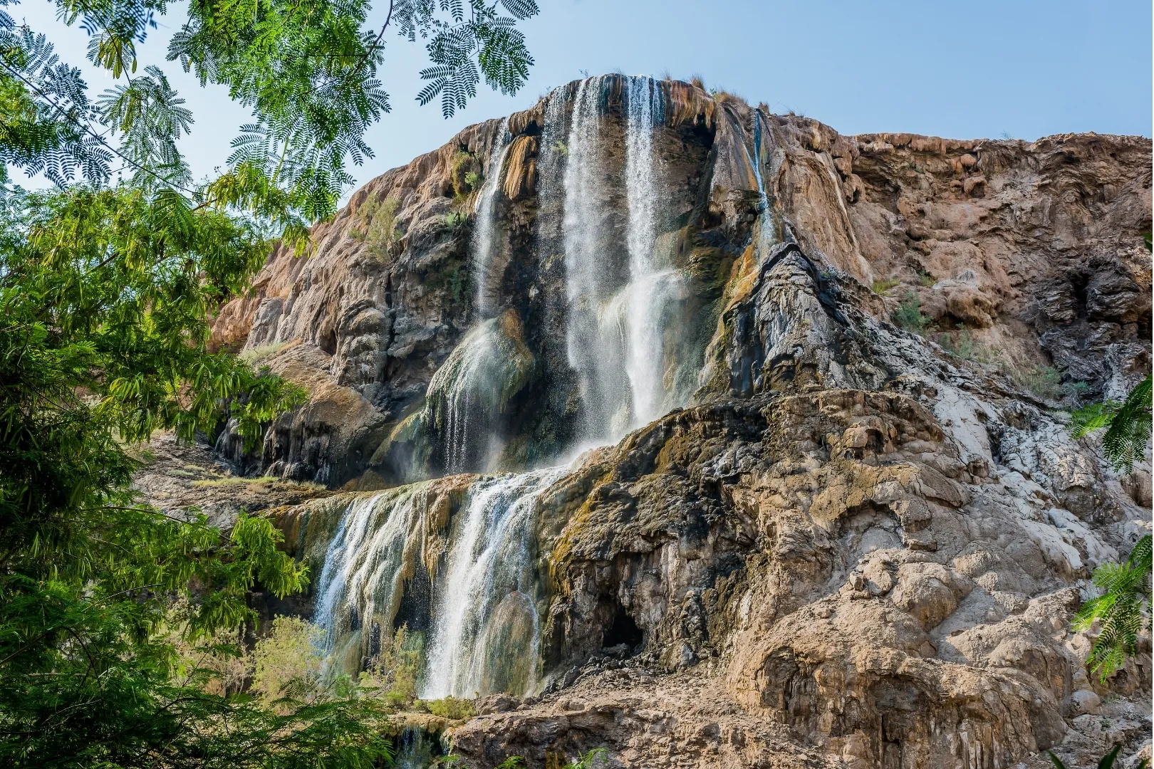 A tall waterfall flowing over layered brown rock at Hammamat Ma'in, surrounded by green tree branches.