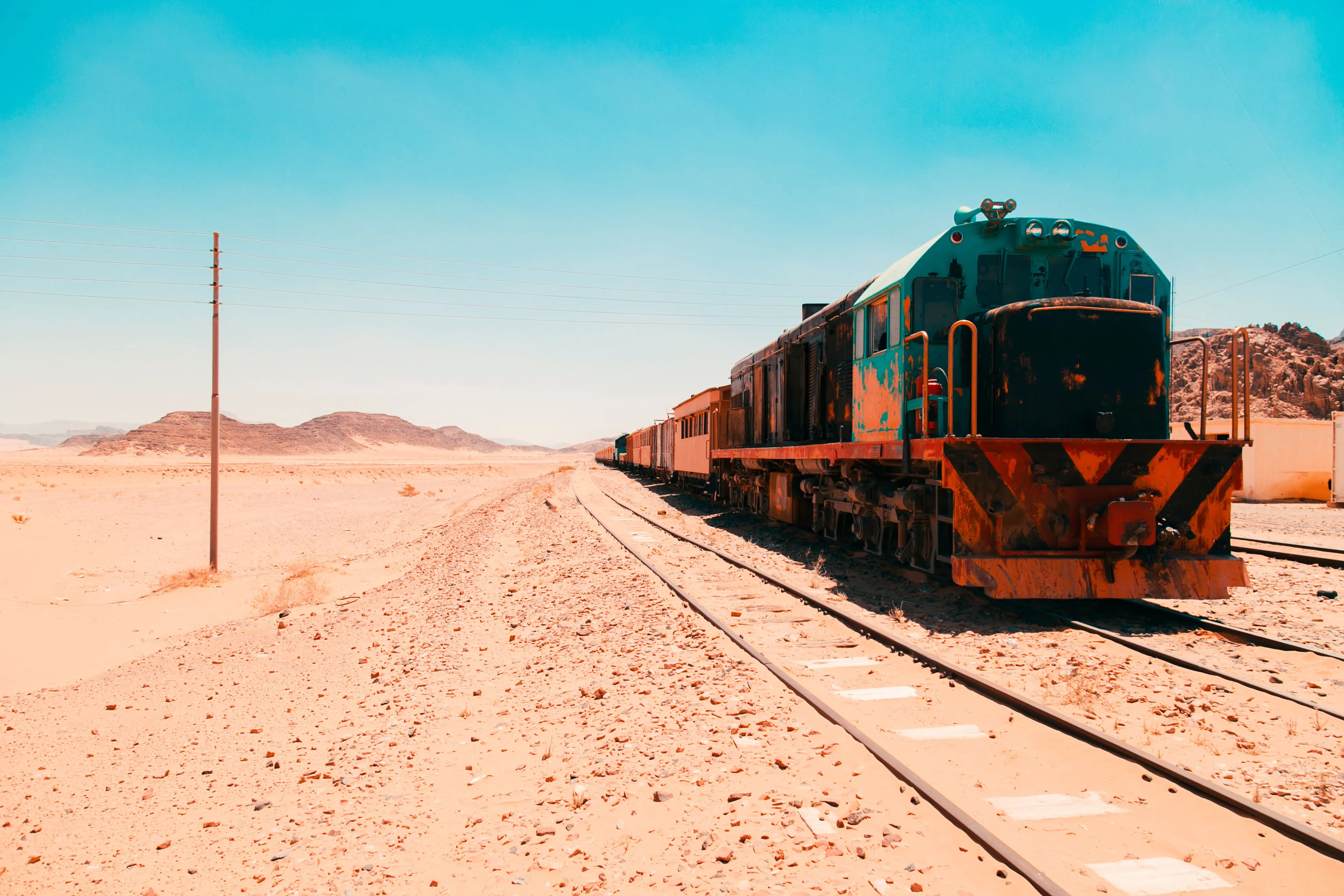 A green and orange train on railway tracks crossing a dry desert landscape beneath a clear blue sky.