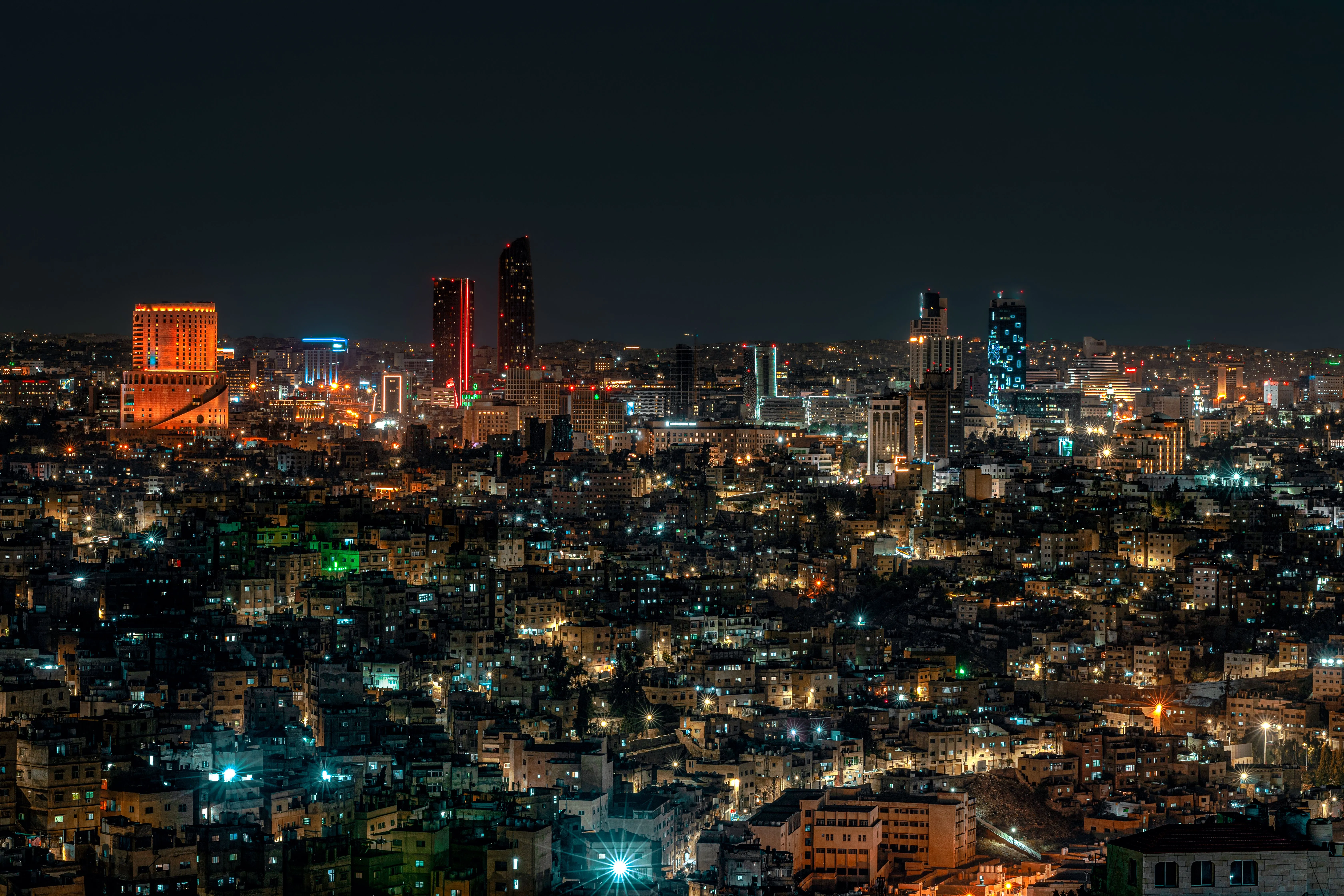 Wide nighttime view of Amman showing densely packed illuminated buildings and several brightly lit modern towers.