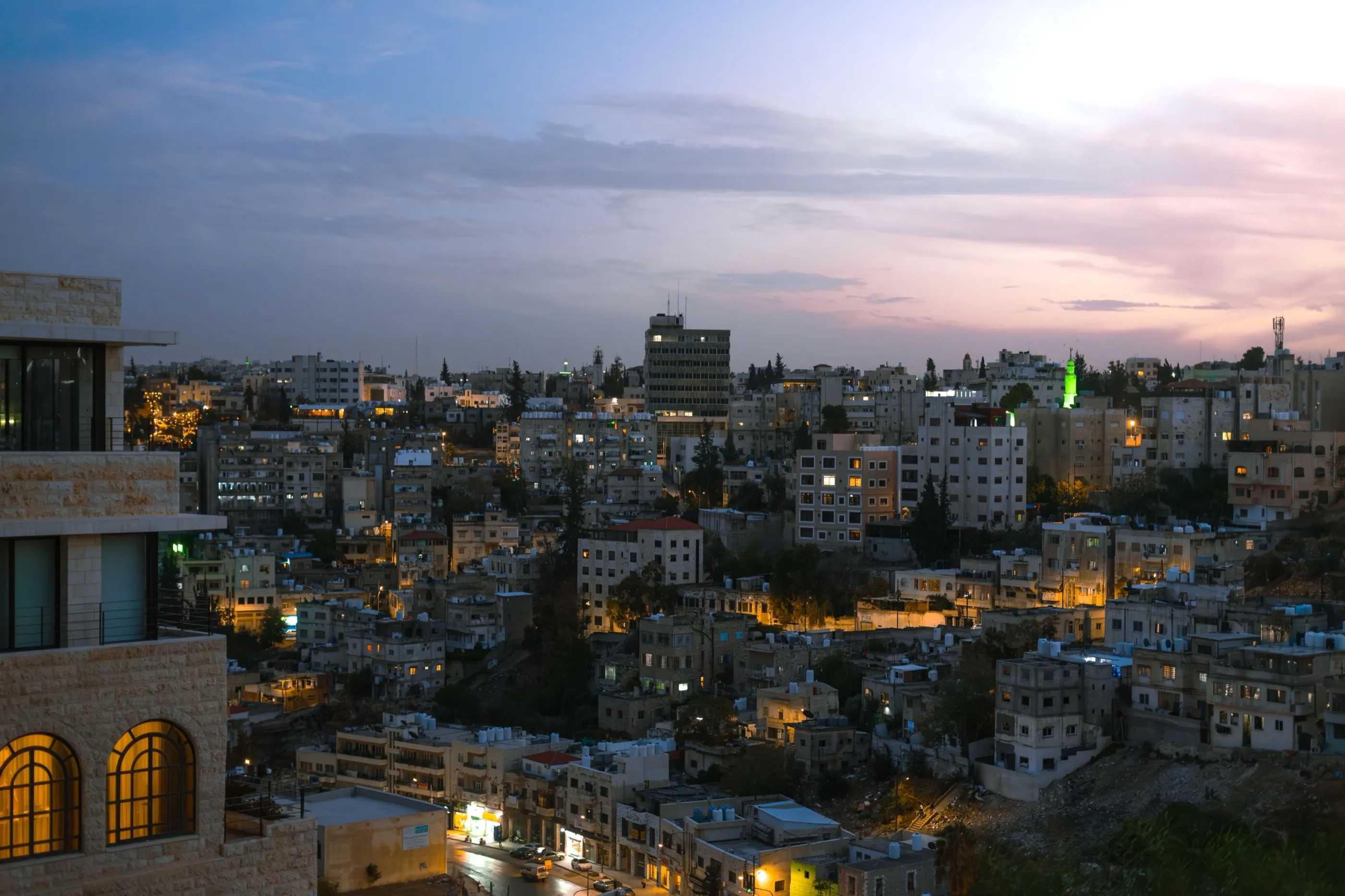 Amman at nightfall, apartment blocks stepping down a hillside with windows and street lights coming on.